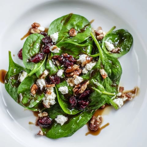 Fresh baby spinach topped with crumbled goat cheese, candied pecans, and dried cranberries, tossed in tangy balsamic vinaigrette in a white bowl.